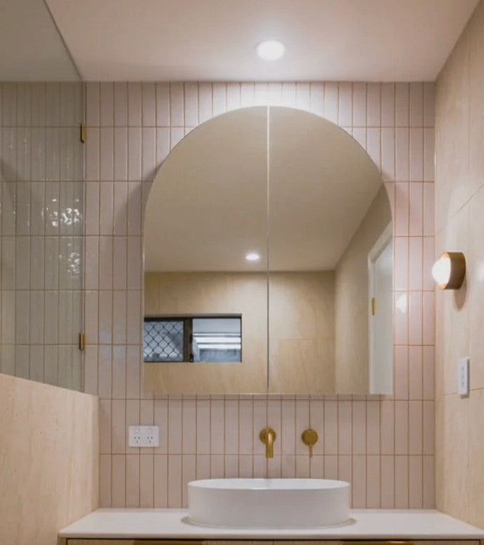 The Eterna Arch mirror cabinet with its doors closed, above a white basin with brass tapware and pale pink vertical tiles